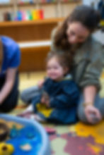 A smiling young child wearing a dark blue smock sits on the floor, enjoying creative play alongside her parent