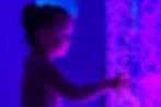 A young girl touches a glowing, bubble-filled tube illuminated with purple and blue lights, her face lit softly