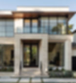 A modern two-story house with large glass windows, a balcony, and a minimalist design. The exterior features light-colored stone, black-framed doors and windows, and a small landscaped garden in front.