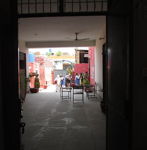 A view through a dark, open doorway into a sunlit courtyard. Several people, mostly women in colorful clothing and headscarves, are gathered in the bright outdoor space. The courtyard features red brick walls, potted plants, and a few chairs, with a ceiling fan visible in the covered walkway leading to it.
