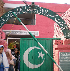 The image shows the entrance to a school building with red brick walls and a green metal gate. Above the gate, a green archway features calligraphy in Urdu script with the school's name. A large Pakistani flag, displaying a white crescent moon and star on a green field, is painted on the gate. A group of young women wearing headscarves stands near the entrance. To the right of the gate, a signboard displays the "School Ranking Board" and the institution's position among 13 total schools.