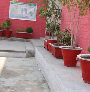 A school courtyard with bright red brick walls and concrete walkways. A row of large red planters containing small trees and shrubs lines a raised ledge. On the wall in the background, a large sign displays a map of the girls school, showing the layout for the 1st and Ground floors.