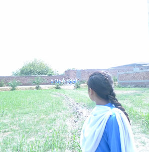 An outdoor shot showing the back of a young girl with a long braid, wearing a blue school uniform and white scarf. She is standing in a green field, looking toward a distant group of similarly dressed students walking along a brick wall. A tall metal tower stands on the right side of the background.