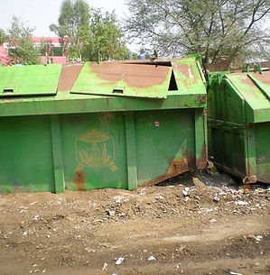 Two large, green metal dumpsters with rusted lids sit on a dirt lot littered with trash. Trees and a red building are visible in the background.