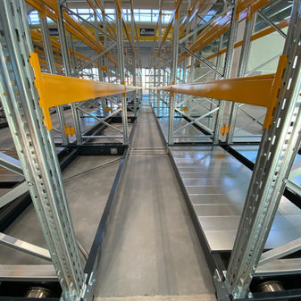 The image shows the interior of a compactible shelving system, with multiple parallel metal racks and walkways creating a visually striking, industrial-style storage environment.