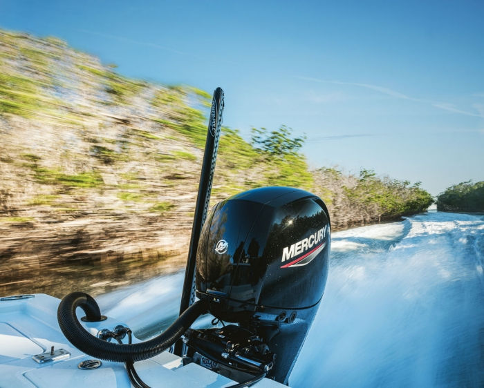 Boat with a Mercury engine speeds through water, creating a trail of spray. Background shows blurred trees under a clear blue sky.