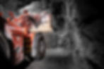 Red mining vehicle with a large scoop maneuvers in a dimly lit rocky tunnel. The rugged, textured walls contrast with the machinery.