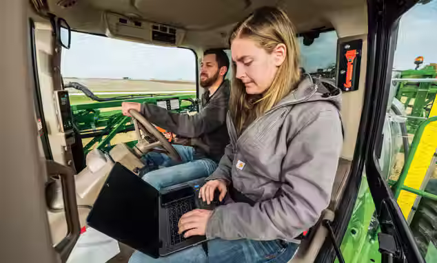 A woman with laptop sitting next to a man that is driving a John Deere Equipment.