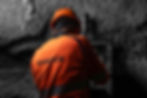 Worker in orange uniform with "SANDVIK" text, wearing a helmet, operates equipment in a dim, rocky underground setting.