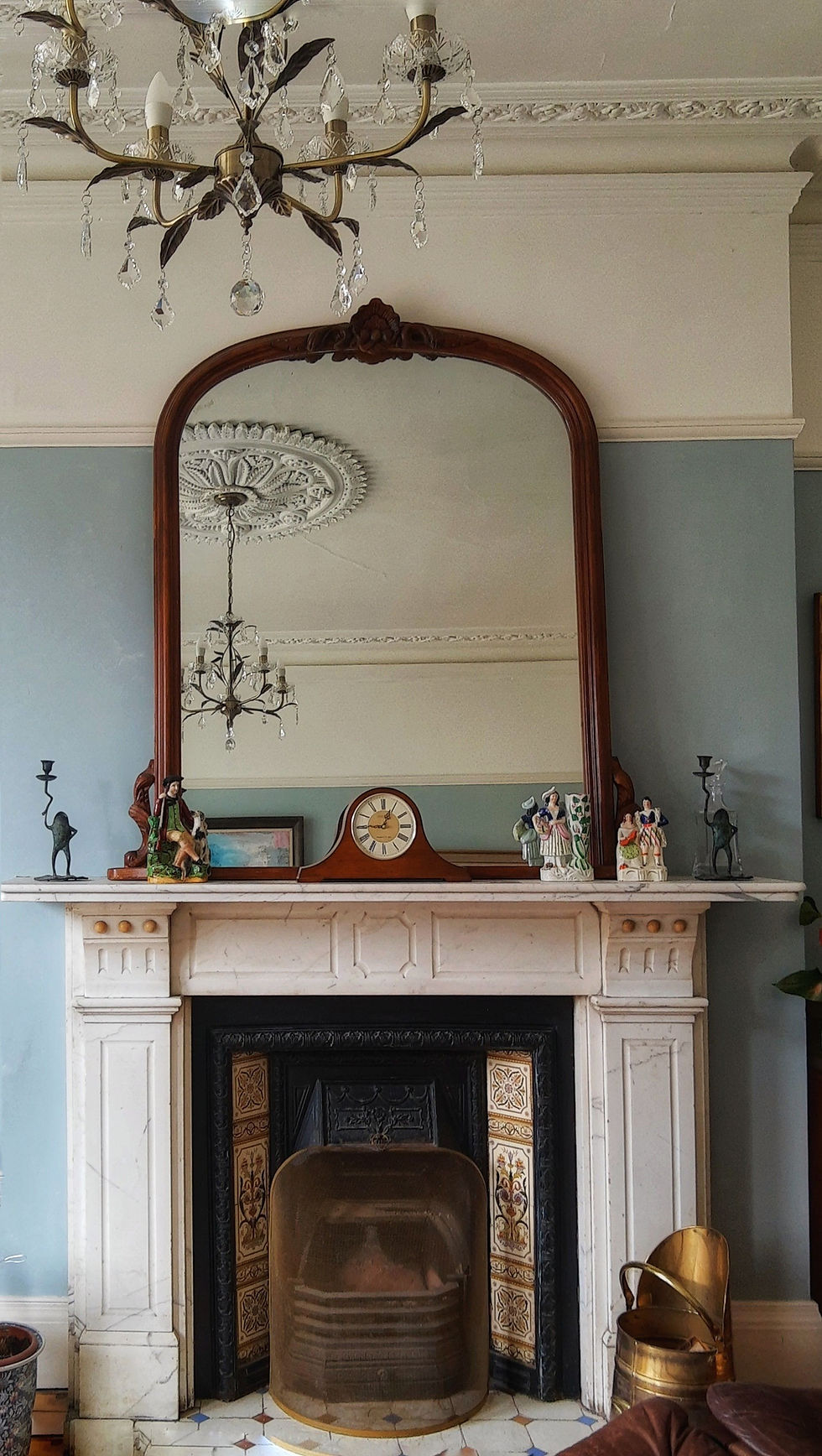 Elegant room with a white marble fireplace, a mirrored mantel, vintage decor, figurines, and a clock. A chandelier reflects in the mirror.