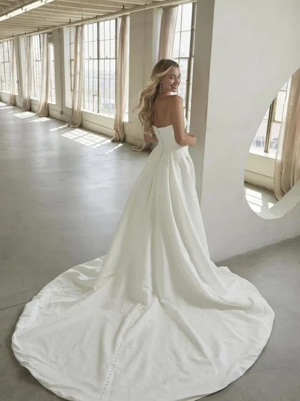 Strapless floral jacquard wedding dress with curved neckline, structured bodice, dramatic front draping, and buttoned train detail.