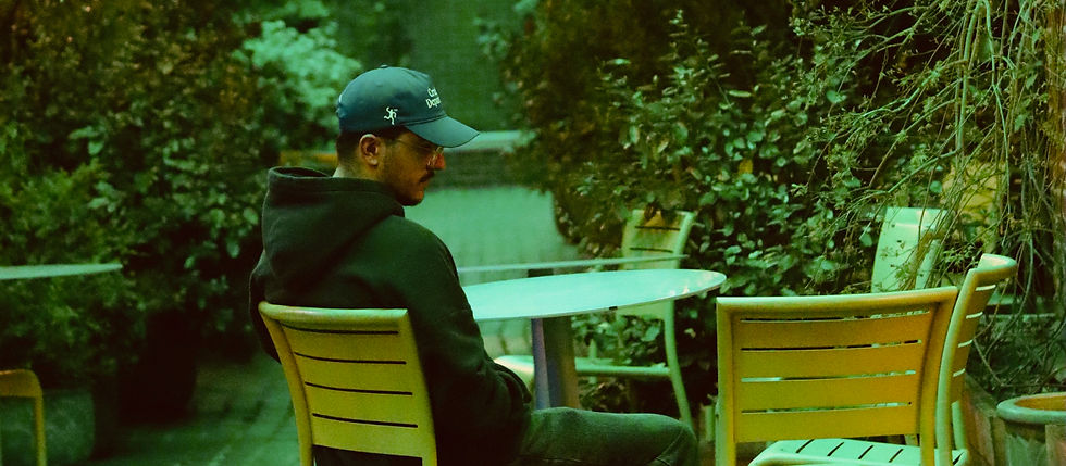 Andrømeda LEFT BEHIND single cover — artist sitting alone at an outdoor table surrounded by greenery