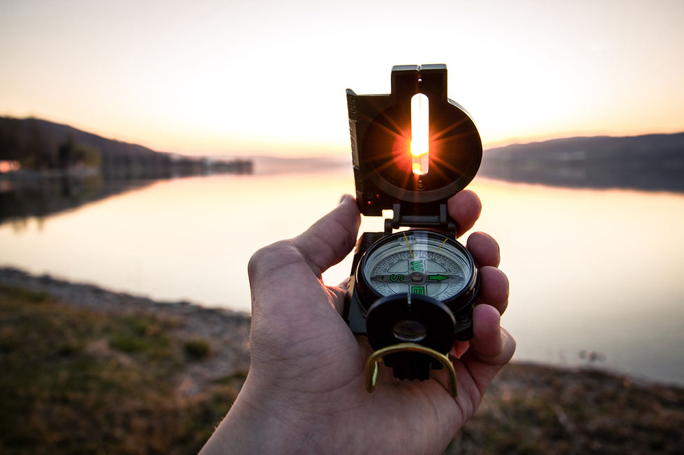 Hand holding a compass against a sunset over a calm lake. The sun shines through the compass’s sight, creating a serene, reflective mood.