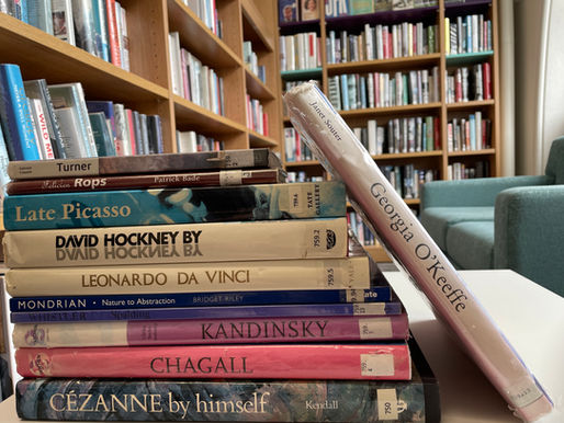 A Pile of art books on a table in a library, 9 on male artists and 1 on a female artist.  Book shelves and a light turquoise sofa in the background.