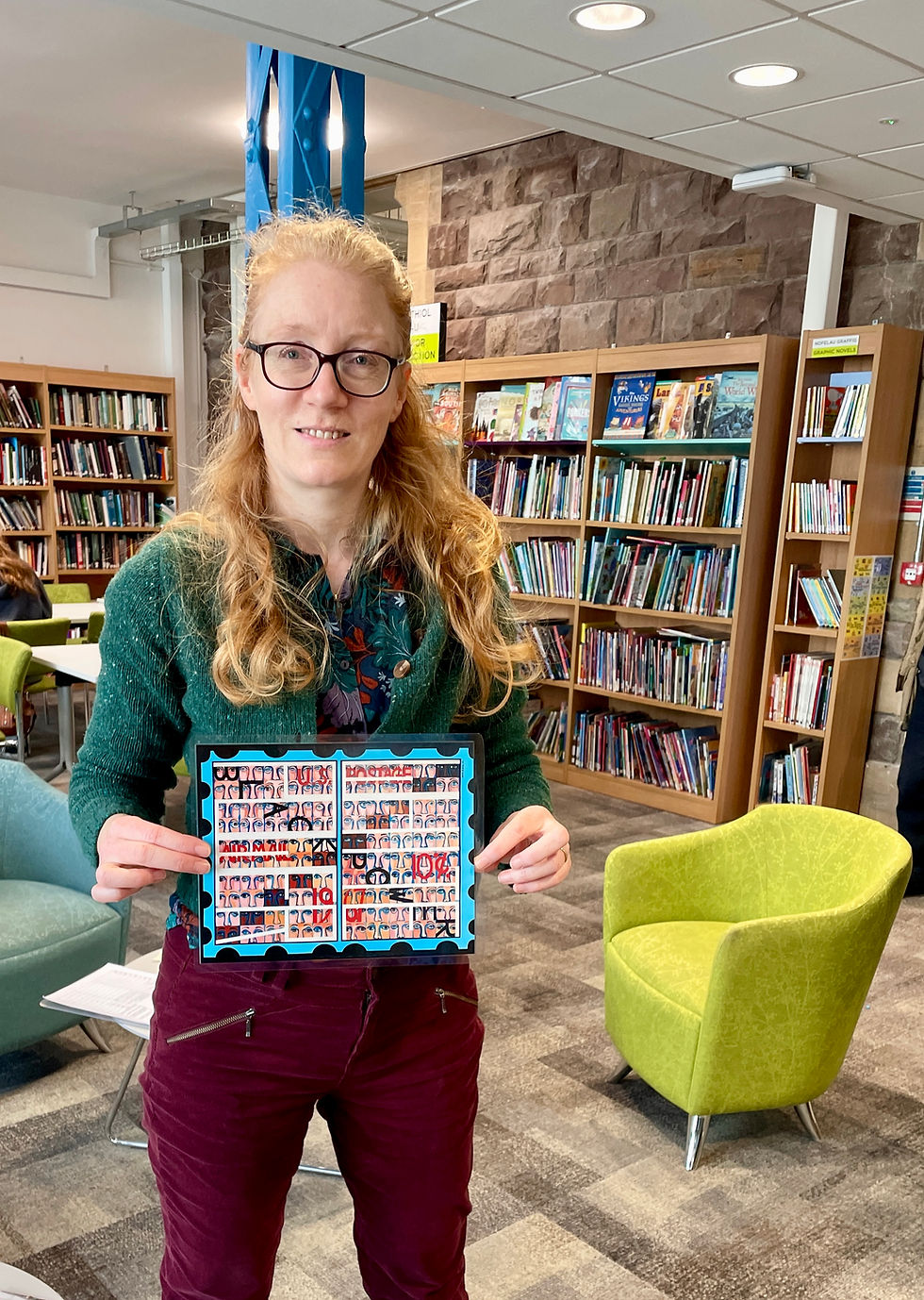 A woman with medium long strawberry blond hair in a library with bookshelves and easy chairs behind her. She is wearing a richly coloured botanical shirt, green cardigan and maroon trousers with zips and holding up an A4 image of a painting by the Black American artist Faith Ringgold. The easy chairs behind are lime green and aqua and you can see Welsh stonework above the book shelves. The carpet is grey tiled effect. The women has brown rimmed glasses and is smiling looking directly at the camera.