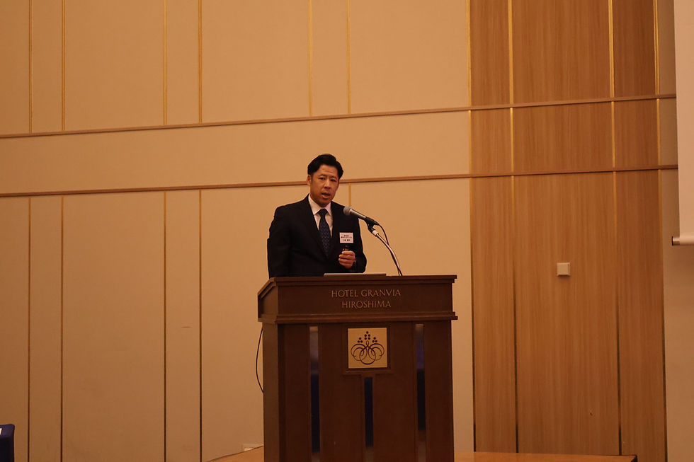 講演中のスーツ姿の社長。背景は木目調の壁で、社長はマイクの前で話している。