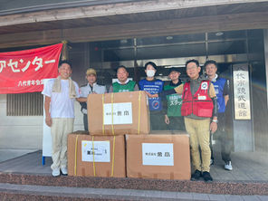 八代市武道館の入口で、笑顔の男性7人が大きな段ボール箱を囲んで立つ。赤い横断幕と施設名の看板が見える。