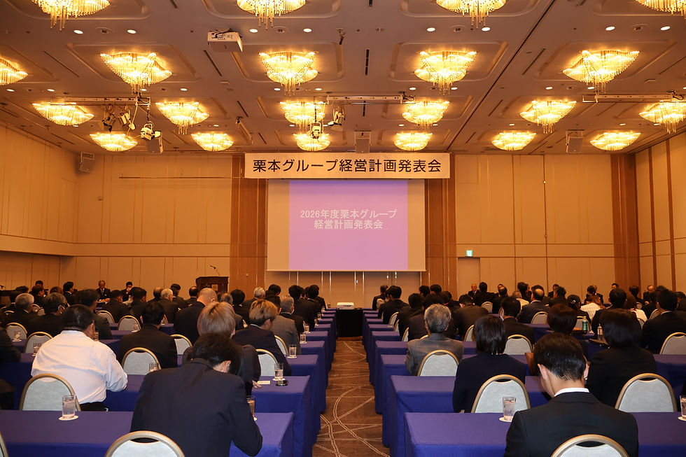 ホテルの会議室で「栗本グループ経営計画発表会」。スーツ姿の参加者が前方のスクリーンを注視。豪華なシャンデリアが輝く。