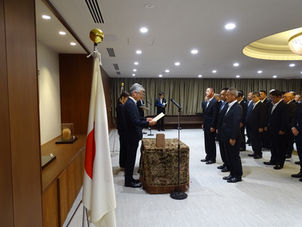 日本国旗の前で、スーツ姿の男性たちが静かに整列し、前方の男性がマイク前で書面を読み上げる式典会場。