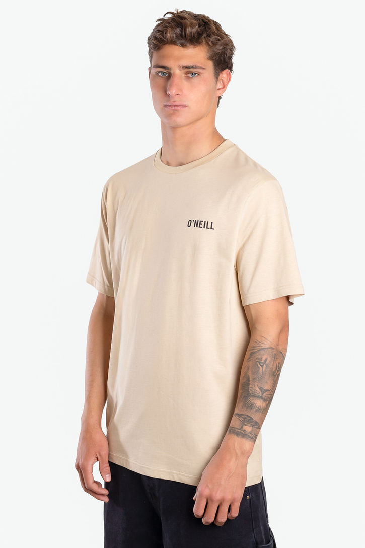 Fotografía de e-commerce de Francisco Doria: un modelo masculino de perfil sosteniendo una pose natural. Viste una remera de algodón de manga corta en color beige claro, con el logo negro de "O'Neill" impreso en el lado izquierdo del pecho. Viste también pantalones oscuros y tiene un tatuaje de león en su antebrazo izquierdo. El fondo es completamente blanco puro. La iluminación es uniforme y resalta la textura de la prenda. Fotografía de catálogo de indumentaria para hombre en Buenos Aires.