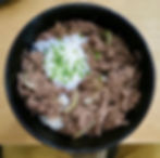 Black bowl with rice topped with sliced beef, onions, a poached egg, and chopped green onions. Placed on a wooden table.