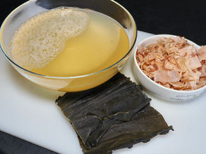 Glass bowl of clear Japanese dashi stock, white bowl with dried bonito flakes, and dried kelp on a white surface, set against a dark background.