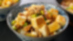Bowl of mapo tofu with tofu cubes, minced meat, and green onions in a patterned bowl, set against a dark background. Warm, appetizing.