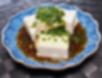 Tofu cubes topped with chopped Shiso herbs sit in a blue patterned bowl with soy dressing, on a dark woven surface. The scene is fresh and appetizing.