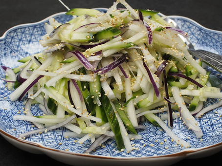 Shredded cucumber and radish daikon salad with sesame seeds on blue patterned plate with silver utensils, set against a dark background.