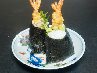 Two shrimp tempura Musubi rice balls on a decorative plate with floral patterns, set against a dark background. The rolls are wrapped in seaweed.