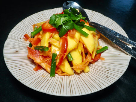 Colorful mango salad with carrots, tomatoes, green beans, and cilantro on a striped plate. Silver utensils on a dark surface.