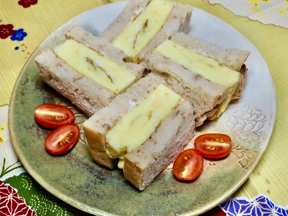 Four egg sandwiches on a decorative plate with halved cherry tomatoes. Background features a colorful, floral tablecloth.