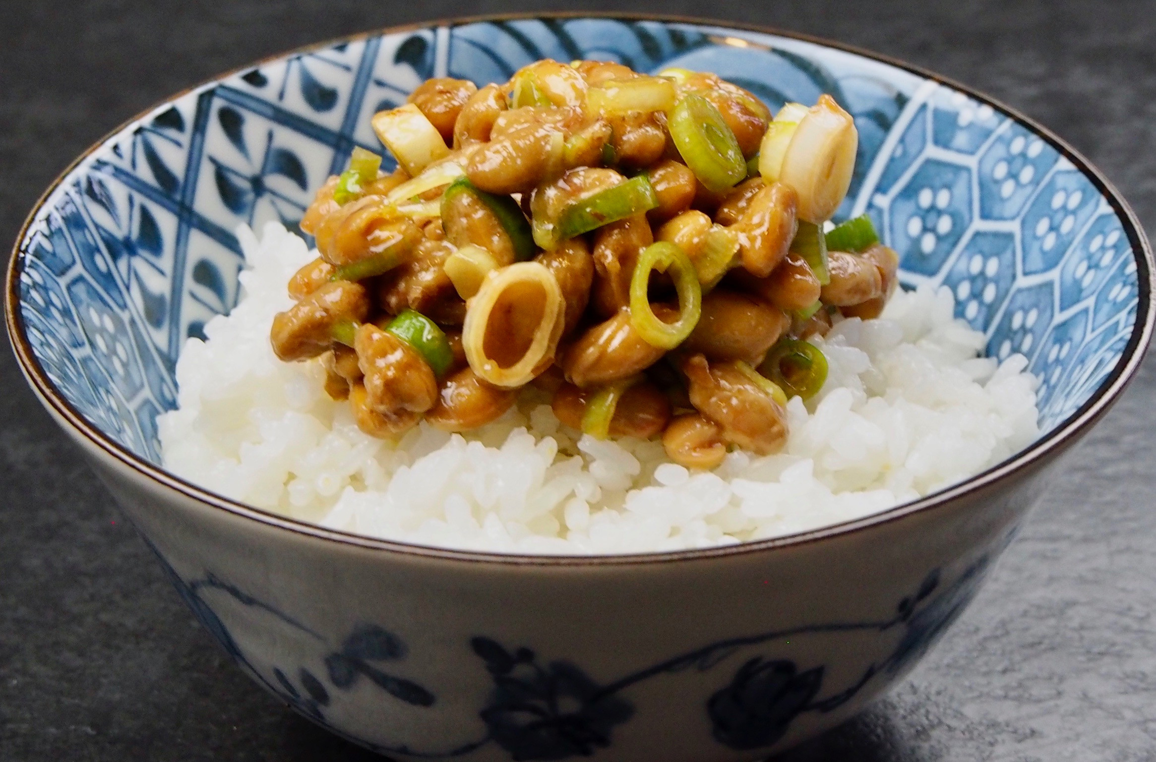 A blue patterned bowl holds white rice topped with natto and sliced green onions, against a dark gray background.