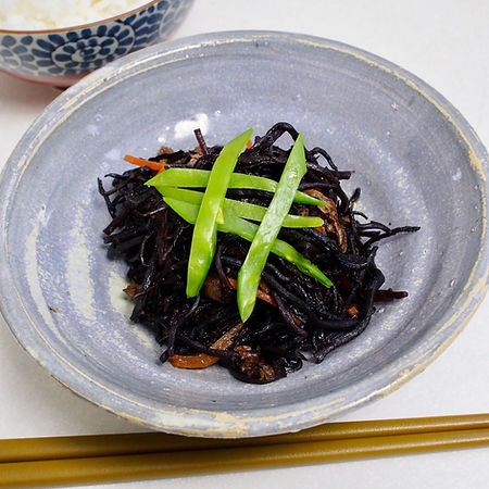 Bowl of simmered hijiki with green beans on top, set on a grey plate. Background includes a patterned bowl and chopsticks, creating a serene dining scene.