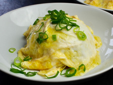 Kimchi Omurice topped with chopped green onions on a white plate. Background shows another similar dish, creating a tasty and inviting scene.