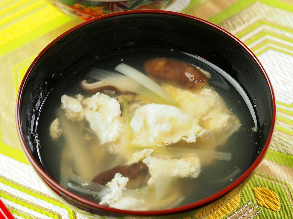 A bowl of clear soup with mushrooms and egg, placed on a vibrant green and yellow patterned cloth. A bowl of rice is in the background.