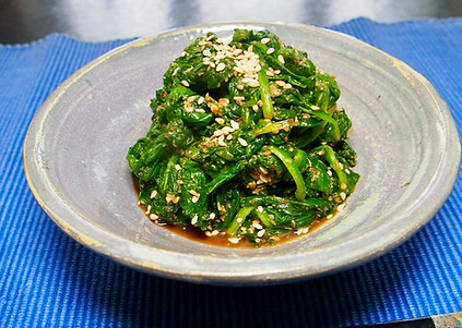 Plate of seasoned spinach topped with sesame seeds, served on a gray dish over a blue placemat. Goma Ae.