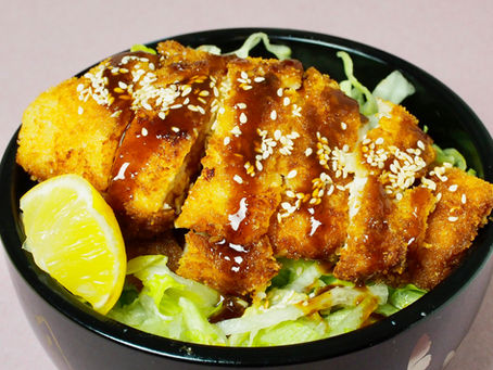 Crispy breaded chicken katsu topped with sesame seeds and sauce on shredded lettuce in a black bowl, with a lemon wedge for garnish.