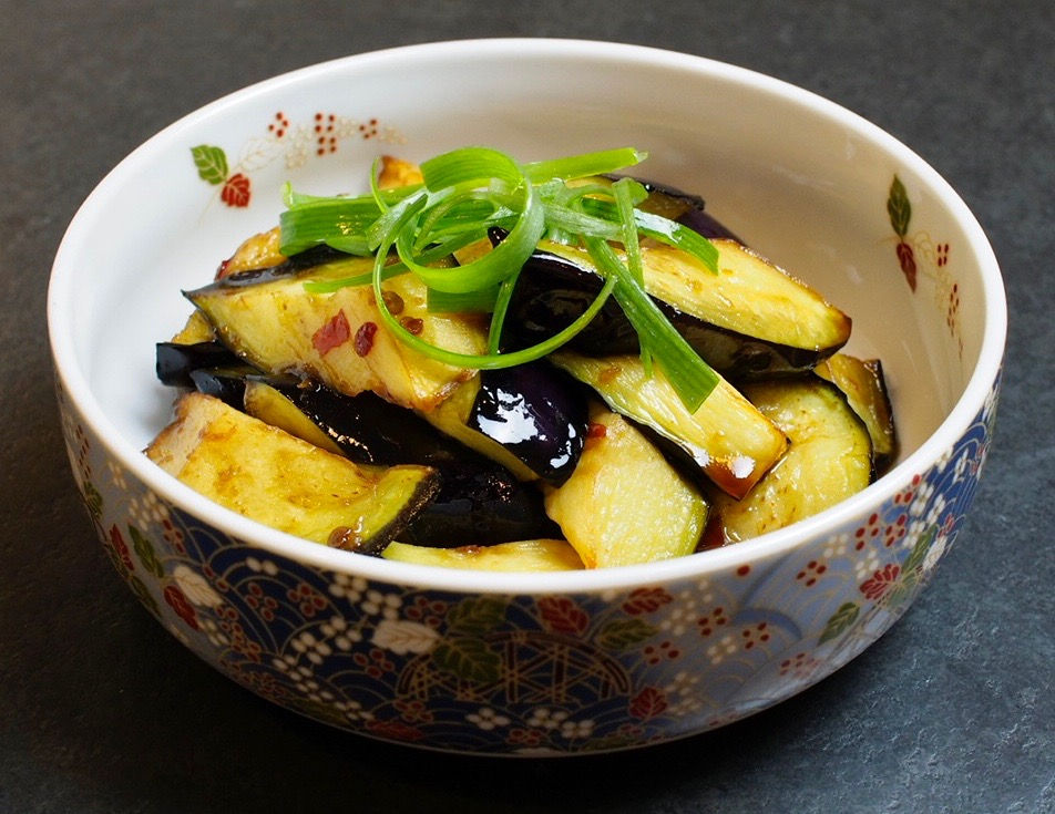 Decorative bowl of glossy Aubergine / eggplant in spicy soy marinade topped with green onion on a dark countertop