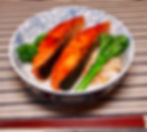 Bowl of teriyaki salmon and broccolini on rice, in a blue floral bowl on a striped cloth. Red chopsticks lie beside the bowl.