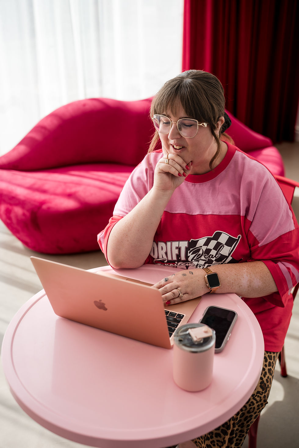 Woman in glasses working on a laptop at a pink table, smiling pensively. Red couch and curtains in the background, relaxed mood.