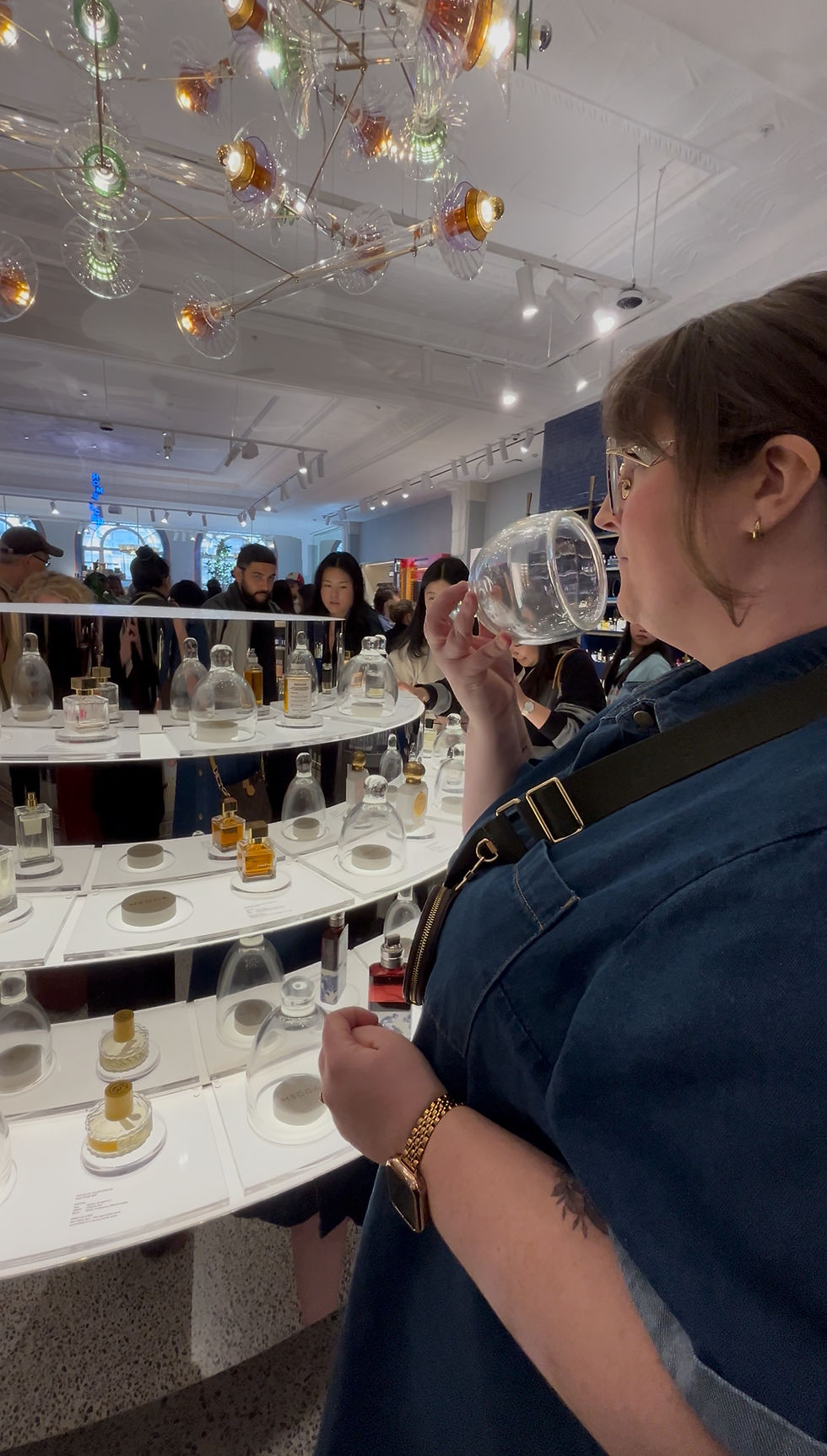 Woman smelling perfume under glass dome in a brightly lit store with a chandelier. People in background observe various bottles.