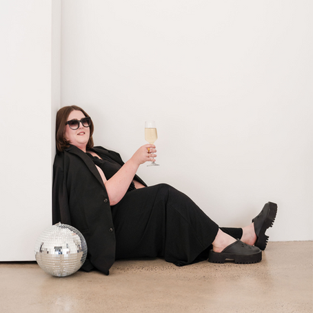 Woman in black outfit, wearing sunglasses, sits on floor holding a wine glass beside a disco ball. White background, relaxed mood.