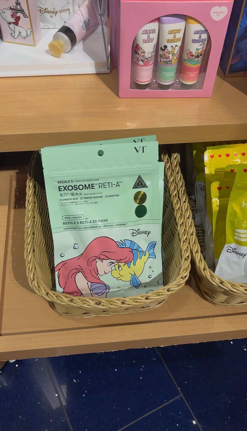 A store shelf with Disney skincare products, featuring Ariel and Flounder on a mint green mask packet in a wicker basket. Bright pink box above.
