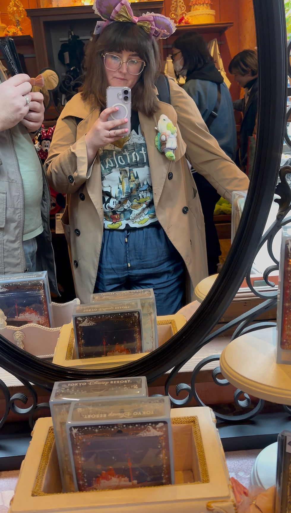 Woman in a trench coat takes a mirror selfie at a shop, wearing large purple ears. Disney-themed shirt, plush toy on coat. Vibrant setting.