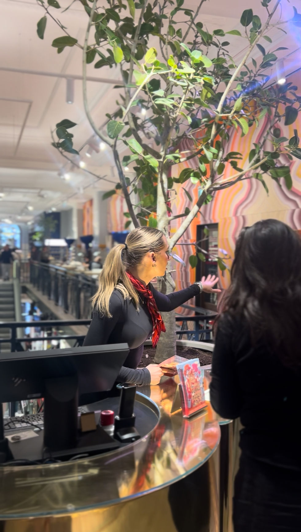 Woman in black, red scarf, gestures at tall plant in stylish store. Vibrant wall pattern. Engaged atmosphere. Customer nearby.