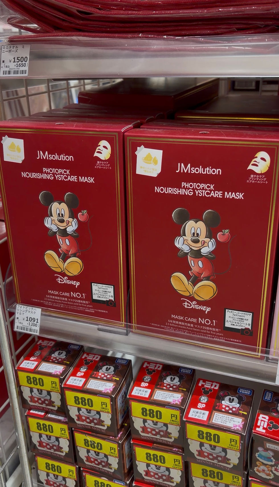 Red boxes of JMsolution Disney face masks featuring Mickey Mouse on a store shelf. Price tags show ¥1500 and ¥1091, ¥880 below.