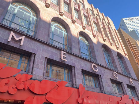 People walk past a red MECCA storefront with bold decorations. A person in a colorful outfit stands by a window. Tall brick building above.