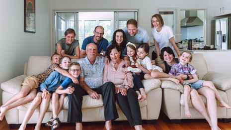 A multigenerational family gathered in a bright living room, with grandparents seated on a sofa surrounded by children and adults standing behind them, all smiling and sharing a warm, candid moment together.
