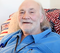 Photographic image of an elderly gentlman with shoulder length gray hair and wearing a blue zip-up shirt is smiling gently while sitting on a recliner covered with a patriotic blanket.
